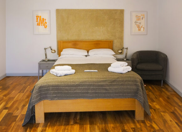 A cozy bedroom with a wooden double bed centered against a beige headboard, neatly made with pillows and a dark quilt. Folded towels are on the bed. A dark upholstered armchair sits to the right, and wall art frames the bed on both sides. The floor is hardwood. B Simply Rooms Stow on The Wold