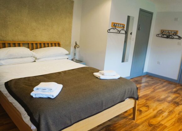 A cozy bedroom features a double bed with white linens and brown throw blanket, two folded towels on the bed, wooden floors, and minimal decor with bedside tables and lamps. A hanger rack and closed door are visible on the light-colored walls. At B Simply Rooms Stow on The Wold