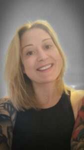 A lady with shoulder-length blonde hair is smiling at the camera. They are wearing a black sleeveless top, showcasing tattoos on their arms. The background is a gradient of gray tones, creating a subtle vignette effect. This is Frankie Copley, the owner of B Simply Rooms Stow on the Wold.